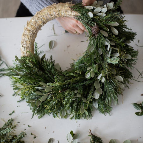 Handmade wreath being crafted on a white surface with greenery.